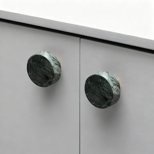 Op de afbeelding: Close-up van twee ronde kastknoppen. De knoppen hebben een groen marmerpatroon en goudkleurige bases. Ze zijn gemonteerd op een lichtgrijze kastdeur. De knoppen geven de kast een vleugje elegantie.