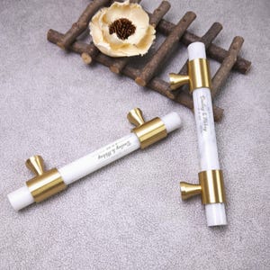 Peut inclure: Deux poignées de placard cylindriques blanches avec des embouts et des supports de montage en métal doré brossé. Chaque poignée porte le texte imprimé Dailey & Abbey 02.05.2025. La quincaillerie est placée près d'une fleur beige clair.