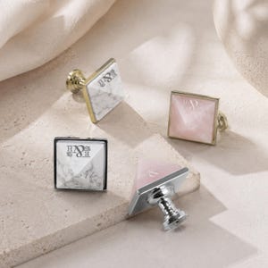 May include: Four square cabinet knobs with pyramid-shaped tops. Two knobs feature a white marble pattern with gold-tone bases, and two feature a smooth pink finish with silver-tone bases. Each knob is engraved with the letters E&H on the top surface.