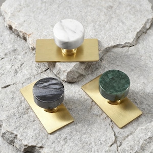 Op de afbeelding: Drie kastknoppen met marmeren toppen in wit, zwart en groen, gemonteerd op rechthoekige messing achterplaten. De knoppen zijn cirkelvormig en zitten op een messing voet. De achtergrond is een getextureerd, lichtgrijs stenen oppervlak.
