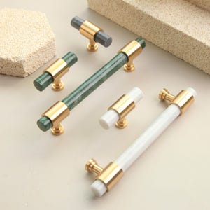 May include: Cabinet hardware set featuring several knobs and pulls. The knobs and pulls have gold-colored bases and accents. The pulls have a long, cylindrical shape with a white or green marble-like center. The knobs have a shorter, cylindrical shape with a gray, green, or white center.