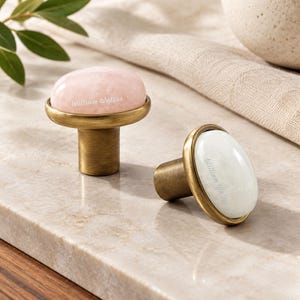 May include: Two cabinet knobs with brushed brass bases and smooth, rounded stone tops. One knob features a pale pink stone, while the other has a white stone. Each stone is engraved with the text William Walker in a delicate, cursive white font.