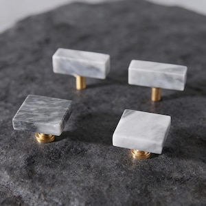 Puede incluir: Cuatro tiradores de mármol para armarios con vástagos de latón. Dos tiradores son rectangulares y dos cuadrados. El mármol es blanco con vetas grises. Los tiradores están sobre una superficie gris oscura texturizada.