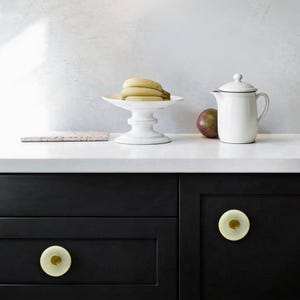 May include: A kitchen scene featuring a white countertop with a white pedestal fruit bowl holding bananas, a white pitcher, and a mango. Black cabinets with gold-toned knobs are in the foreground. A small, rectangular, pinkish-tan coaster is also present.