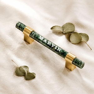 May include: A cylindrical cabinet handle featuring a dark green marbled pattern with gold-toned metal mounting brackets at each end. The name MARVIN is printed in white capital letters along the centre of the green handle surface.