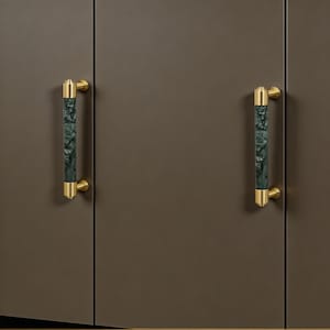 May include: Two cabinet door handles. Each handle has a cylindrical, dark green and gray marbled center with gold-colored end caps and mounting hardware. The handles are mounted on a brown cabinet door.