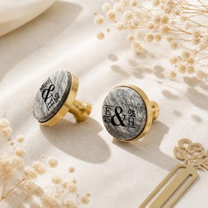 Op de afbeelding: Twee ronde kastknoppen met een goudkleurige metalen voet en een ronde bovenkant met een grijs en wit marmerpatroon. Elke knop is bedrukt met de zwarte tekst E & H in een decoratief schreeflettertype, gecentreerd op het marmeroppervlak.