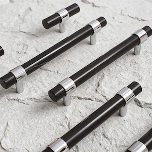 May include: A collection of cabinet pulls featuring a cylindrical black bar design with polished chrome metal accents at each end and mounting posts. The hardware displays a sleek, modern aesthetic with visible branding on the chrome collar.