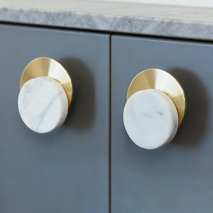 May include: Close-up of two cabinet doors with decorative hardware. Each door has a gold-toned circular backplate with a white marble knob. The cabinet doors are a muted grey colour, and the countertop is a light grey.