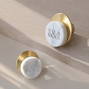 May include: Two circular cabinet knobs featuring a white marble face with the engraved letters E & H and decorative flourishes. Each knob has a brushed gold metal base that extends beyond the marble surface, creating a layered, circular profile.