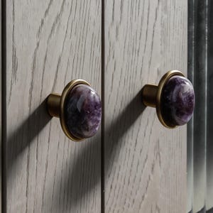 May include: Two cabinet knobs with oval, purple gemstone centers and brass-colored metal rings and bases. The knobs are mounted on a light gray wooden cabinet door.