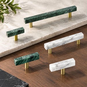 May include: A collection of rectangular cabinet pulls and knobs featuring green or white marble bars with gold-toned metal posts. Each piece is engraved with the text MARVIN. The set includes two long bar pulls and two smaller T-bar knobs.