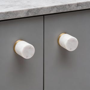 Op de afbeelding: Close-up van twee kastknoppen. Elke knop heeft een witte marmeren cilinder met grijze adering, geplaatst op een goudkleurige basis. De knoppen zijn gemonteerd op een grijze kastdeur, met een marmeren aanrechtblad op de achtergrond.