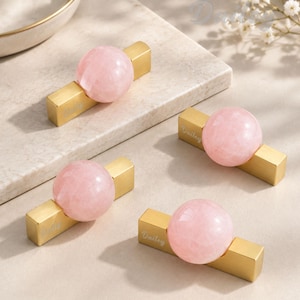 May include: Four drawer pulls featuring a spherical light pink stone knob attached to a rectangular brushed gold metal base. Each gold base is engraved with the word Dailey in a script font. The design combines a smooth, round gemstone element with a structured, metallic rectangular component for furniture hardware.