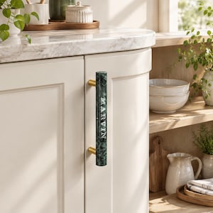 May include: A cylindrical cabinet handle made of dark green marble with white veining, featuring the name MARVIN engraved in white capital letters. The handle is mounted vertically on a cream-colored cabinet door with two gold-toned metal brackets.