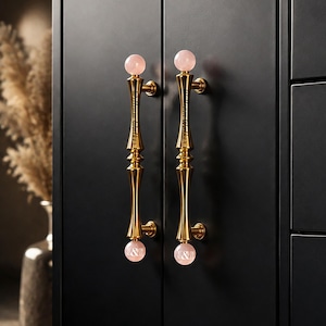 May include: Two vertical gold-toned metal cabinet handles mounted on a matte black door. Each handle features a polished gold finish with a slender, contoured central shaft and a spherical light pink stone finial at both the top and bottom ends.
