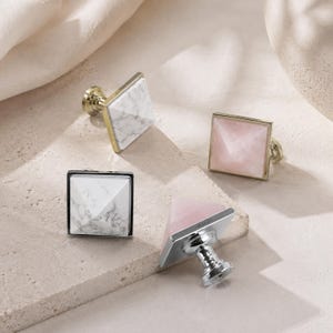 May include: Four square cabinet knobs with pyramid-shaped tops. Two knobs feature white marble with grey veining and gold-toned bases, while two knobs feature translucent pale pink stone with silver-toned bases. Each knob has a central metal post.