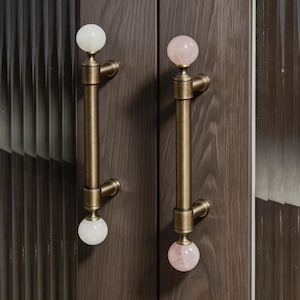 May include: Two cabinet pulls with a vintage brass finish. One pull has white stone spheres, the other has pink stone spheres. The pulls are mounted on a dark wood cabinet door.