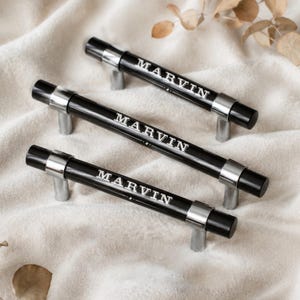 Peut inclure: Trois poignées de placard cylindriques avec une finition noire brillante et des supports de montage en métal argenté. Chaque poignée est gravée du nom MARVIN dans une police serif, accompagnée de petites étoiles de chaque côté du texte.