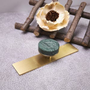 May include: A green marble circular knob with the text VALENTIR centred on a rectangular brushed brass plate. The knob features a smooth, polished finish with natural white veining, while the brass base has a flat, matte metallic surface.