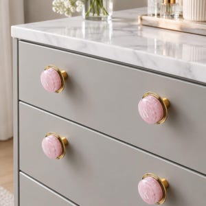Puede incluir: Cuatro pomos circulares para armarios instalados en una cómoda gris. Cada pomo presenta una base de metal dorado pulido y una inserción de resina rosa claro translúcida y lisa con un sutil símbolo de ampersand blanco en el centro.