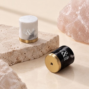 Könnte beinhalten: Zwei zylindrische Möbelknöpfe, einer aus weißem und einer aus schwarzem Marmor, jeweils mit einem goldfarbenen Metallsockel und dem weißen Monogramm E&H. Der schwarze Knopf zeigt einen Gewindeeinsatz an seiner flachen Basis.