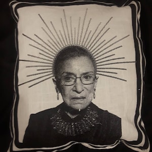 May include: A white pillow with a black border features a black and white image of a woman wearing glasses and a necklace. The woman is surrounded by a radiating sunburst design.
