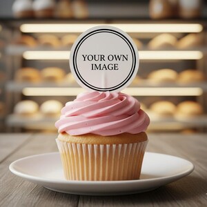 May include: A cupcake with pink icing and a round topper that reads "YOUR OWN IMAGE" sits on a white plate. The cupcake is on a wooden table, with a bakery display case in the background.