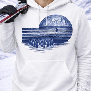 Może przedstawiać: Biała bluza z kapturem z niebieską grafiką przedstawiającą pingwiny, górę i narciarza. Grafika jest stylizowaną ilustracją o okrągłym kształcie. Bluza ma przednią kieszeń i kaptur ze sznurkiem. Tło to zaśnieżony krajobraz.