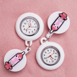 Puede incluir: Dos relojes de bolsillo blancos para enfermeras con un dije de jeringa. Los relojes tienen un movimiento de cuarzo y una esfera blanca con números negros y agujas rojas. Los dijes de jeringa son rosas y tienen una cara linda.