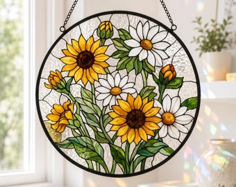 May include: A circular stained glass suncatcher featuring a floral arrangement of yellow sunflowers and white daisies with green leaves. The design is enclosed in a black metal frame and suspended by a thin black chain, set against a clear textured glass background.