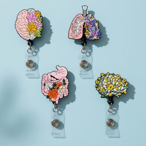 以下が含まれることがあります： 4つの医療をテーマにしたリトラクタブルバッジホルダー。デザインには脳、肺、腸が含まれており、それぞれにカラフルな花のアクセントが施されています。バッジリールは、金属クリップ付きの透明なプラスチックホルダーに取り付けられています。