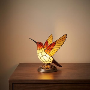 May include: A stained glass table lamp shaped like a hummingbird in flight. The bird features a gradient of orange, yellow, and cream-colored glass panels held together by dark metal lines. It rests on a circular base with a leaf-like design.
