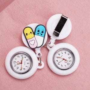 Op de afbeelding: Twee witte verpleegster zakhorloges met quartz uurwerk. Eén horloge heeft een bedel met twee stripfiguur pil karakter. De horloges hebben een witte wijzerplaat met zwarte cijfers en rode secondewijzers. De achtergrond is roze.