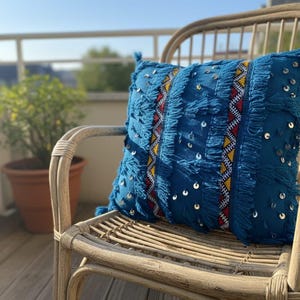 Puede incluir: Un cojín azul decorativo con flecos y lentejuelas descansa sobre una silla de mimbre. El cojín presenta un patrón geométrico con detalles rojos, amarillos y blancos. Una planta en maceta está en el fondo.