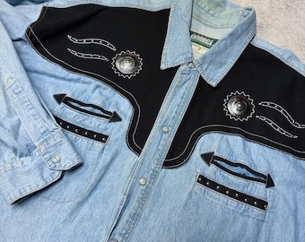 May include: A light blue denim long-sleeve shirt featuring black fabric yokes on the chest and back. The shirt includes silver-toned metal concho buttons, decorative silver studs, and arrow-shaped black trim accents on the two front chest pockets.