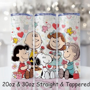 Könnte beinhalten: Drei Edelstahlbecher mit einem Cartoon-Design mit Snoopy, Charlie Brown, Lucy und anderen Peanuts-Charakteren. Die Becher haben einen weißen Hintergrund mit Herzen, Blumen und Vögeln. Der Text unten lautet "20oz & 30oz Straight & Tapered."