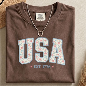 Puede incluir: Una camiseta marrón de manga corta con el texto USA en una fuente universitaria, relleno con un estampado floral. Debajo del texto aparece la frase EST. 1776 con dos pequeñas estrellas. Un collar de cadena plateada descansa en el cuello.