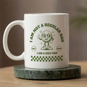 Puede incluir: Taza de cerámica blanca con un diseño verde con temática de golf. El diseño incluye un personaje de pelota de golf con gafas de sol y el texto "I AM NOT A REGULAR DAD I AM A GOLF DAD". La taza está sobre un posavasos de mármol verde.