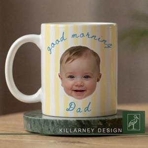Op de afbeelding: Witte keramische mok met een geel en wit gestreept patroon. De mok heeft een lachend babygezicht en de woorden "good morning Dad" in blauw schrift. De mok staat op een groene marmeren onderzetter.