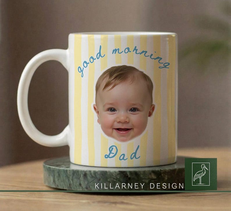 Op de afbeelding: Witte keramische mok met een geel en wit gestreept patroon. De mok heeft een lachend babygezicht en de woorden "good morning Dad" in blauw schrift. De mok staat op een groene marmeren onderzetter.