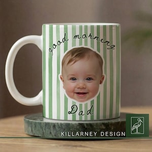 Op de afbeelding: Witte keramische mok met een groen en wit gestreept patroon. De mok toont het lachende gezicht van een baby en de woorden "good morning Dad" in een scriptlettertype. De mok staat op een groene marmeren onderzetter.