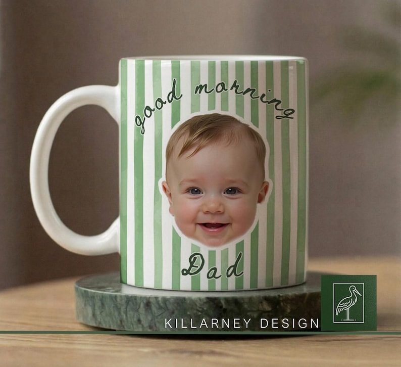 Op de afbeelding: Witte keramische mok met een groen en wit gestreept patroon. De mok toont het lachende gezicht van een baby en de woorden "good morning Dad" in een scriptlettertype. De mok staat op een groene marmeren onderzetter.