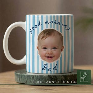 Op de afbeelding: Witte keramische mok met blauwe verticale strepen en de woorden "good morning Dad" in script. Het gezicht van een lachende baby staat in het midden van de mok. De mok staat op een groene marmeren onderzetter.