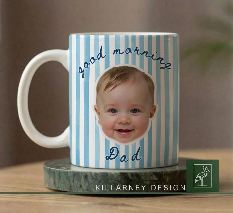 Op de afbeelding: Witte keramische mok met blauwe verticale strepen en de woorden "good morning Dad" in script. Het gezicht van een lachende baby staat in het midden van de mok. De mok staat op een groene marmeren onderzetter.