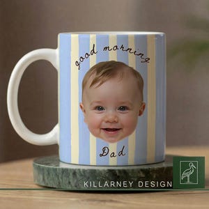 Op de afbeelding: Een keramische mok met een babygezicht en de tekst "good morning Dad" in script. De mok heeft een lichtblauwe en gele gestreepte achtergrond. De mok staat op een groene marmeren onderzetter. De mok heeft een witte handgreep.