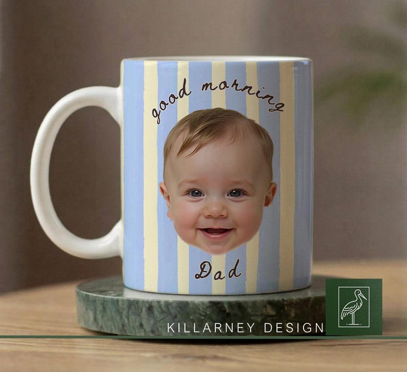 Op de afbeelding: Een keramische mok met een babygezicht en de tekst "good morning Dad" in script. De mok heeft een lichtblauwe en gele gestreepte achtergrond. De mok staat op een groene marmeren onderzetter. De mok heeft een witte handgreep.