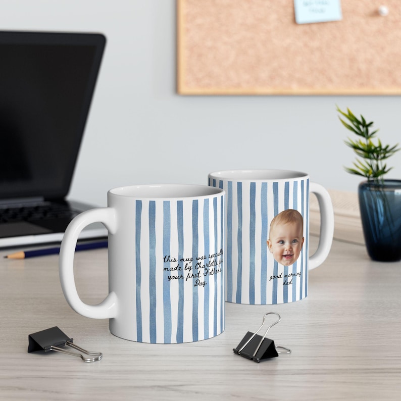 Op de afbeelding: Twee witte keramische mokken met blauwe verticale strepen. E&eacute;n mok heeft een babygezicht en de woorden "good morning dad". De andere mok heeft de tekst "this mug was specially made by Charlotte for your first Father's Day."