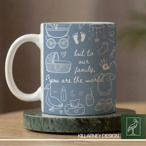 Puede incluir: Una taza de cerámica con asa blanca y fondo azul claro, decorada con dibujos lineales blancos de artículos para bebés y el texto "but to our family, you are the world." La taza está sobre un posavasos de mármol verde.