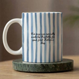 Op de afbeelding: Witte keramische mok met blauwe verticale strepen. De mok heeft een wit handvat en bevat de tekst: "this mug was specially made by Charlotte for your first Father's Day."